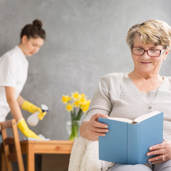 A senior woman reading a book while a woman works on cleaning a table behind her